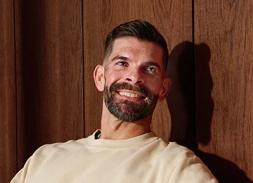 A man with a beard wearing a beige shirt and smiling